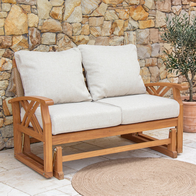 A wooden teak outdoor glider loveseat with two cream cushions, set against a stone wall and potted plant.