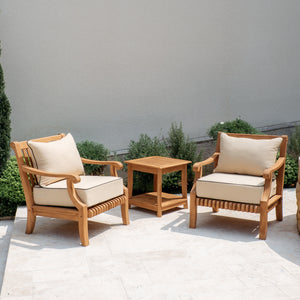 A 3-piece teak outdoor patio furniture set featuring two wooden armchairs with cream cushions and a matching wooden side table, arranged on a light stone patio against a neutral gray wall.