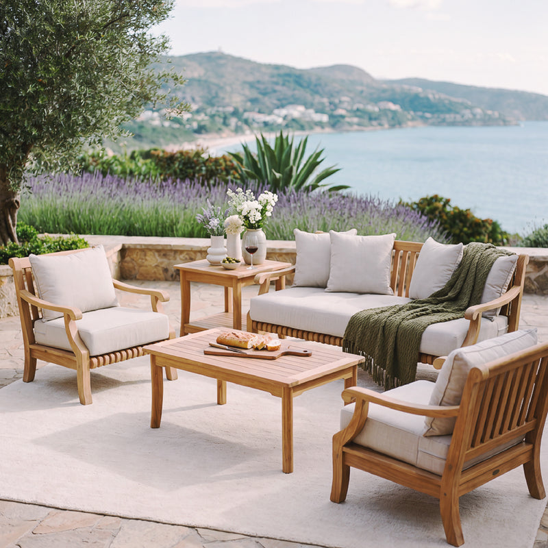Teak outdoor sofa conversation set styled with cushions and a green throw blanket on a coastal patio overlooking the ocean in a serene outdoor setting.