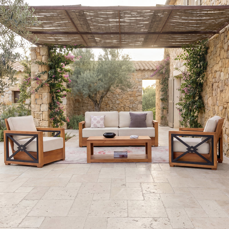 Outdoor patio furniture set in a stone courtyard under a woven roof, featuring a wooden three-seat sofa with cream cushions, two matching armchairs with black X-frame accents, and a rectangular wooden coffee table.